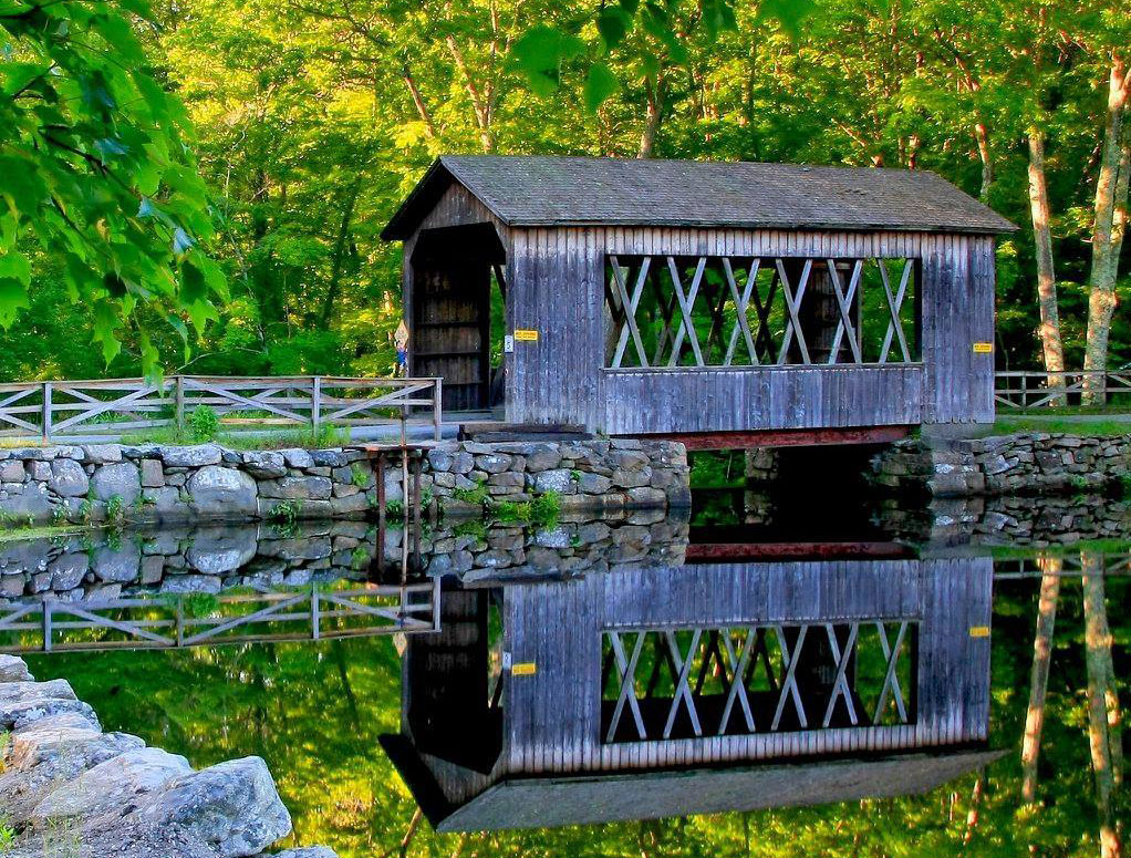Covered bridge at Sutton Falls Campground reflected in calm water on a summer day